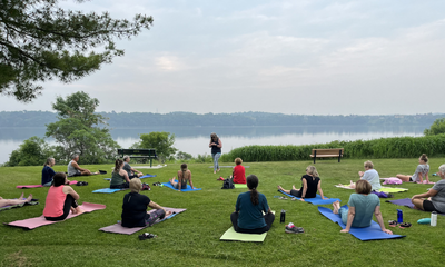 People doing yoga on grass near a lake with a woman instructing.