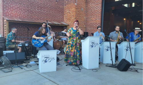 A band performs outdoors in front of a brick wall, featuring a singer and musicians with guitars, keyboards, and brass instruments.
