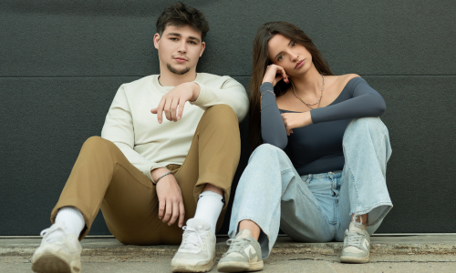 Two people sitting against a dark gray wall in casual clothing.