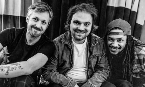 "Three smiling men seated on a couch in a black and white photo."