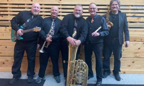 Five men in black attire holding musical instruments, standing against a wooden plank wall.